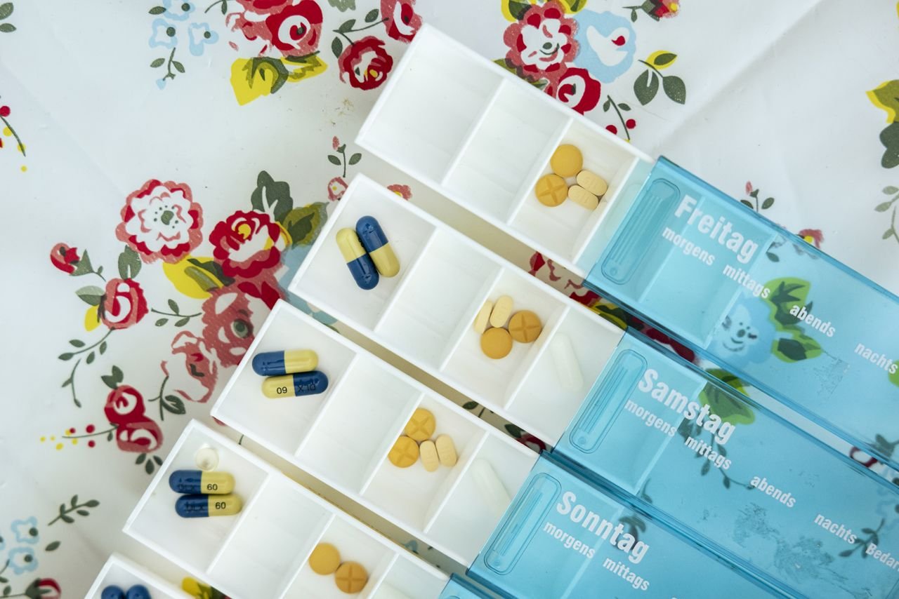 Drei blaue Medikamentenboxen für Freitag, Samstag und Sonntag mit jeweils fünf Fächern für morgens, mittags, abends, nachts und Bedarf. Die Fächer sind unterschiedlich mit Tabletten und Kapseln in verschiedenen Farben gefüllt. Die Boxen liegen auf einer geblümten Tischdecke mit roten, gelben und blauen Blumen.