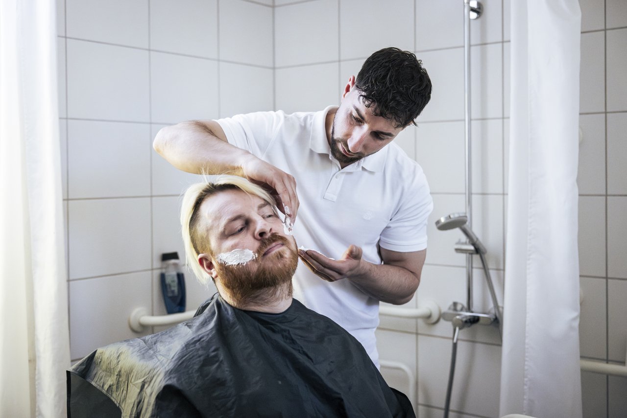 Ein Pfleger rasiert einem Mann im Badezimmer das Gesicht. Der Mann sitzt mit geschlossenen Augen und wirkt entspannt.